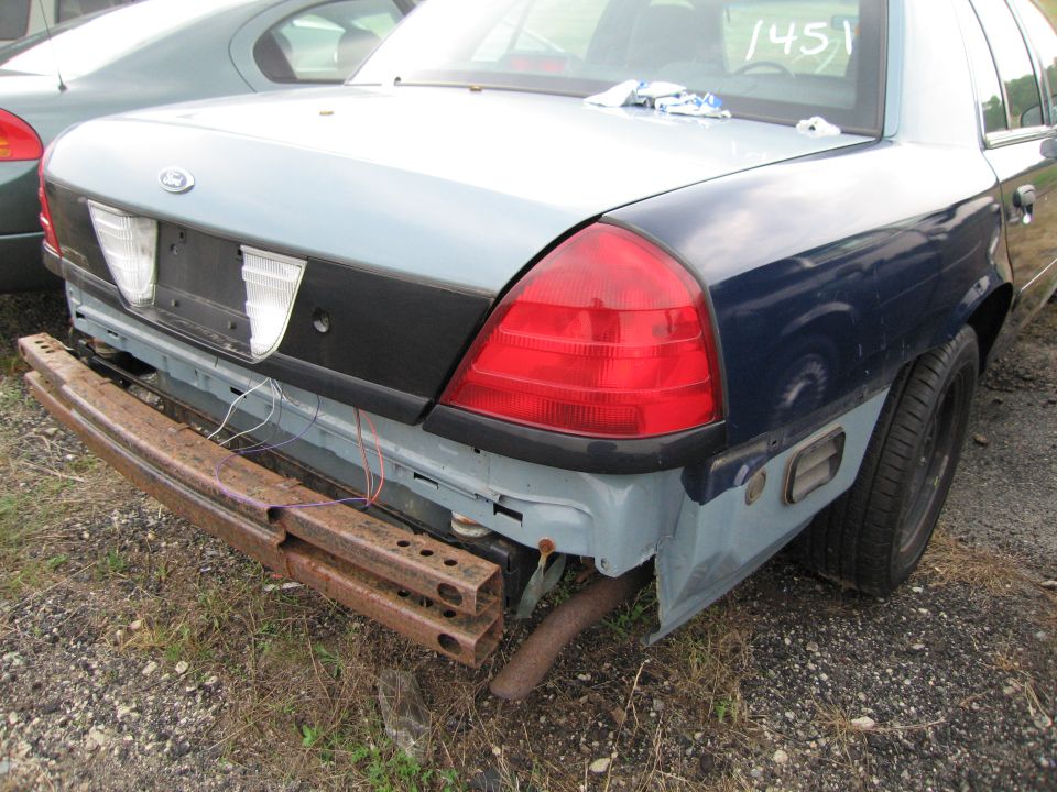 Rear of a Ford Crown Victoria with a rusty aftermarket bumper attached. Another car is parked behind it