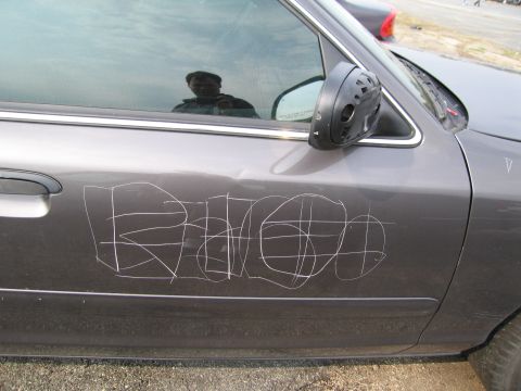 1991 Ford LTD Crown Victoria - The side of a car has been vandalized with white scratches forming graffiti on the door panel.