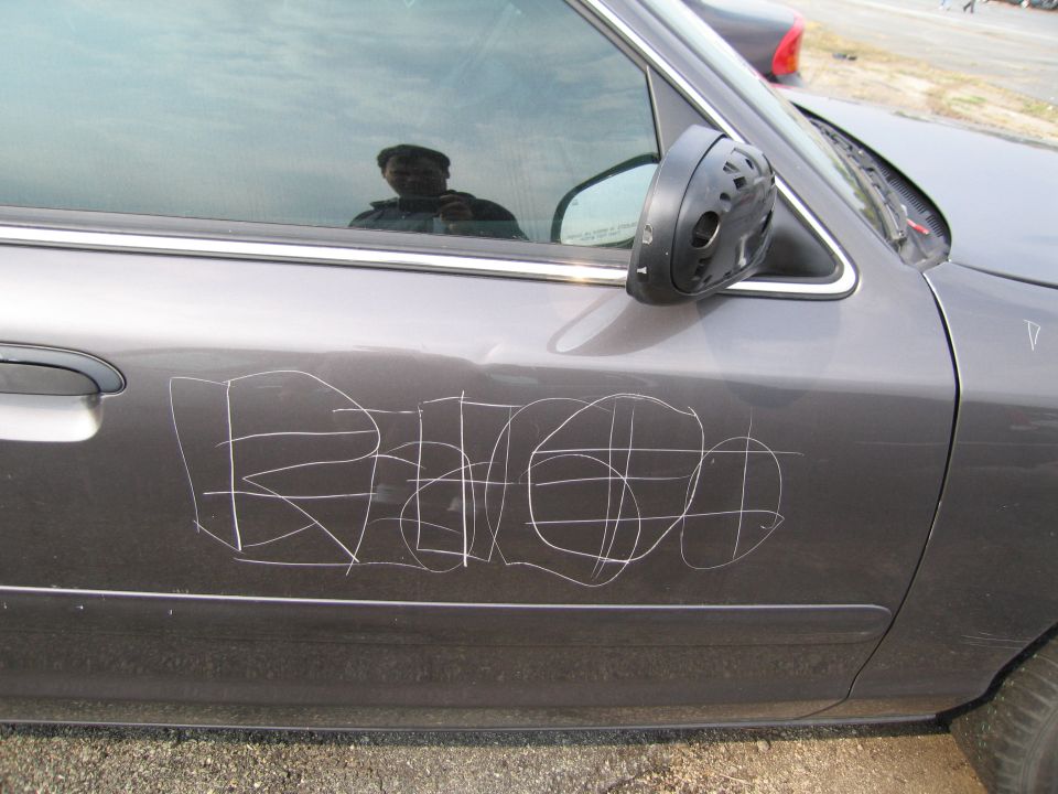 Gray car door with white graffiti scratched into it. Side mirror is visible