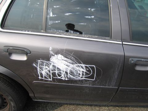 1991 Ford LTD Crown Victoria - A gray sedan's side door is covered in white graffiti and numerous scratches.