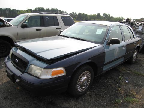 1991 Ford LTD Crown Victoria - A two-tone Ford Crown Victoria sedan sits in a junkyard with its hood open and a damaged front fender.