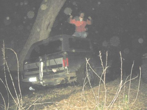 1995 Ford Bronco - A person stands atop a dark SUV, which is parked near a tree in a wooded area at night.