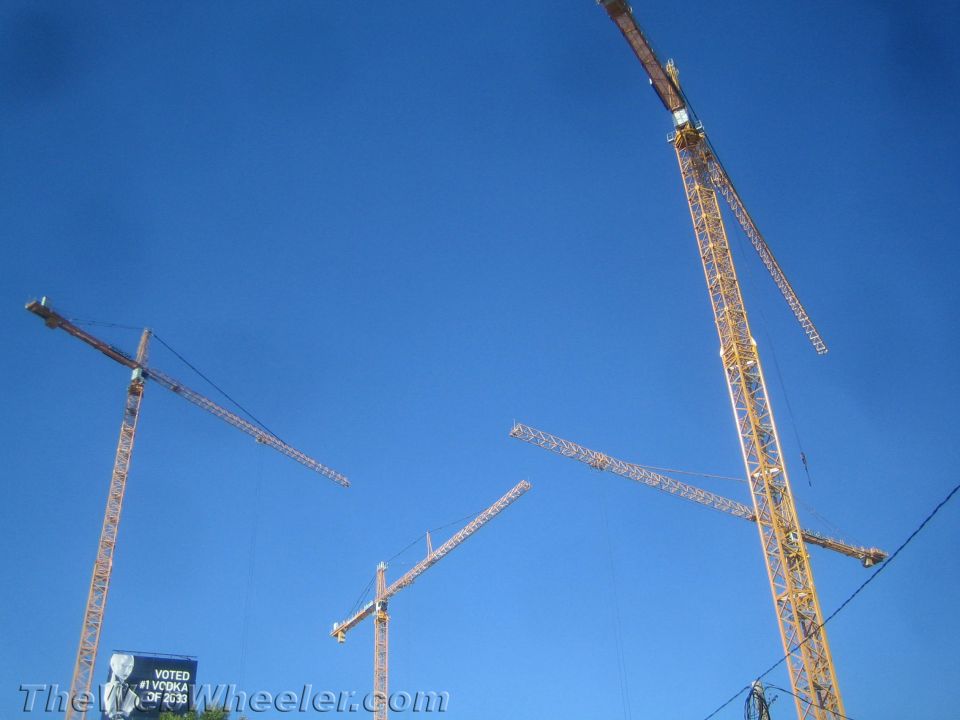 Four yellow construction cranes against a bright blue sky. A billboard is visible in the lower left