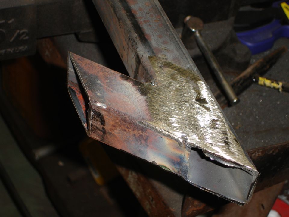 Metal tube with an angled cut and visible weld marks, resting on a workbench with tools in the background