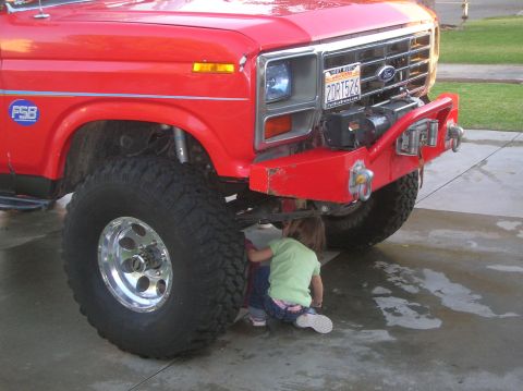 1985 Ford Bronco - Kids check out the underside of a lifted red Ford Bronco, which appears to be from the early 1980s.