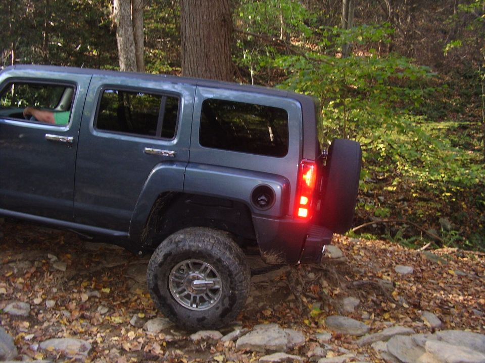 Dark blue Hummer H3 driving on a rocky, wooded trail. Rear tire and taillight are visible