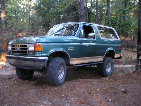 1989 Ford Bronco - Driver-side three-quarter front view of a lifted green and tan Ford Bronco with a tan removable hardtop, parked on pine needles in a wooded…