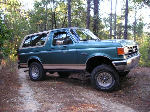 1989 Ford Bronco - Green full-size Ford Bronco with tan removable hardtop and tan lower two-tone flexing on a rutted dirt trail through a pine forest, shot fr…