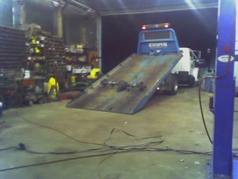 1992 Ford Bronco - A tow truck is parked in a garage, with a white truck loaded onto its flatbed. Tools and shelves are visible in the background.