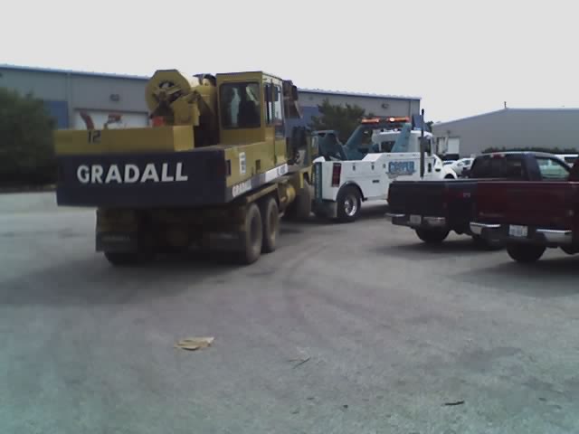 Yellow Gradall machine on truck chassis, rear view. White tow truck and red pickup trucks visible