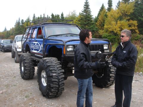 1996 Ford Bronco - Two men converse next to a heavily modified blue SUV in a line of vehicles.