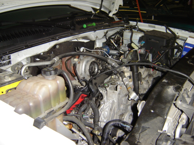 Engine bay with coolant reservoir, hoses, and wiring visible. Some text on a blue box and a red tool
