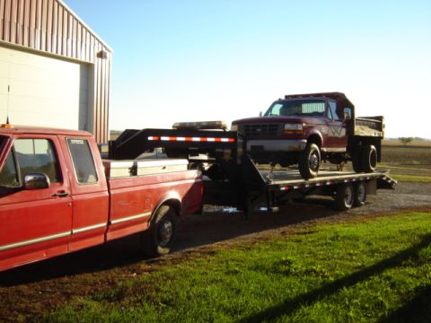 1992 Ford Bronco - A maroon Ford F-350 dump truck is transported on a trailer, with a red pickup truck visible in the foreground.