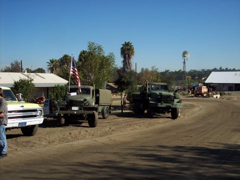 1981 Kenworth W900A - Two olive drab military trucks are parked on a dirt road with buildings and a windmill in the background.