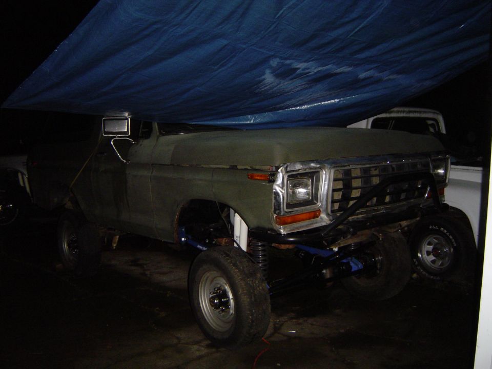 Modified pickup truck with custom front bumper and lifted suspension, partially covered by a blue tarp. Another white truck is visible behind it