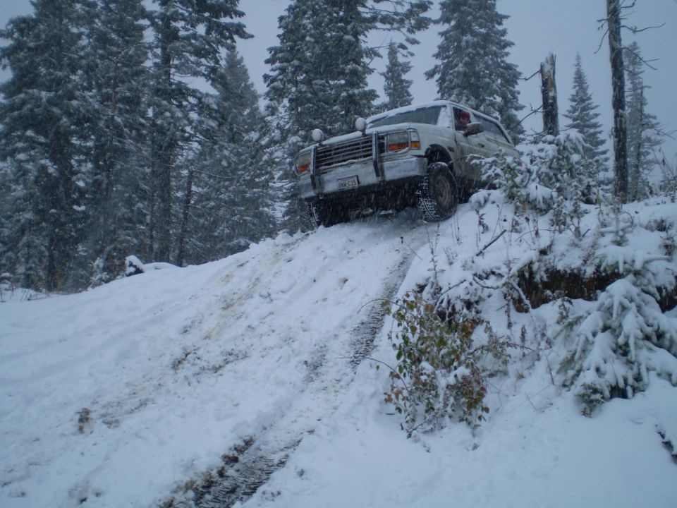 White 1992 Ford Bronco driving up a snowy hill with trees in the background