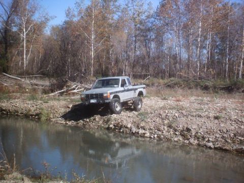 1990 Ford Ranger - A lifted pickup truck is parked on a rocky bank next to a body of water with trees in the background.