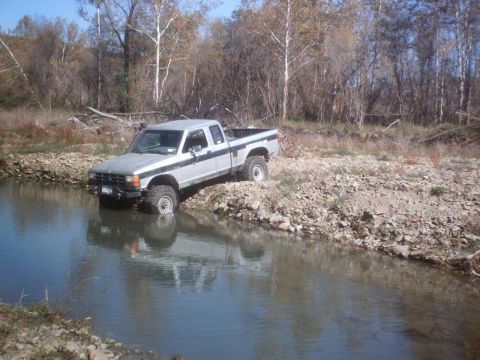 1990 Ford Ranger - A lifted pickup truck navigates a rocky stream bed, its tires partially submerged in the water.