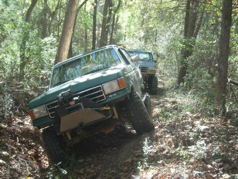 1995 Ford Bronco - Two Ford Broncos navigate a muddy, wooded trail, one equipped with a winch.