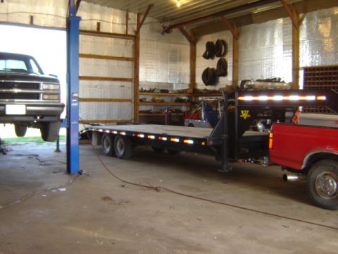 1992 Ford Bronco - A black truck is elevated on a lift inside a garage, next to a large trailer and a red truck.