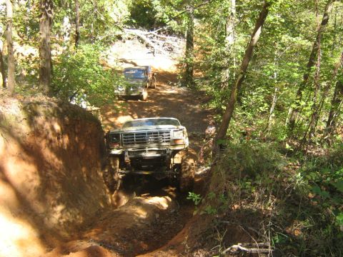 1986 Ford Bronco - Two off-road vehicles navigate a steep, muddy trail through a wooded area.