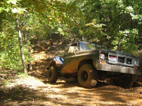 1986 Ford Bronco - A heavily mud-splattered 1977 GMC truck navigates a steep, dirt trail through a wooded area.