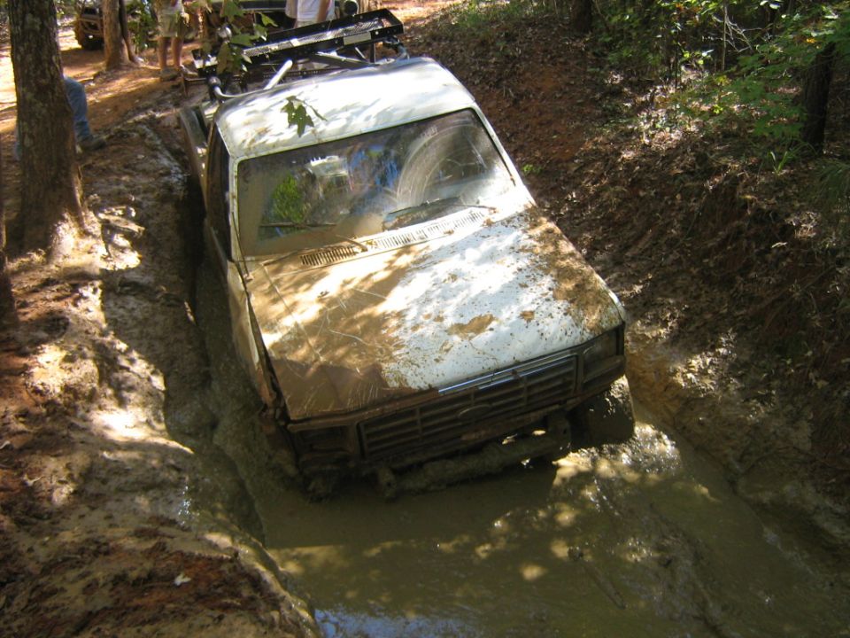 White off-road truck stuck in a muddy ditch, with trees and people around. Another vehicle is visible in the background