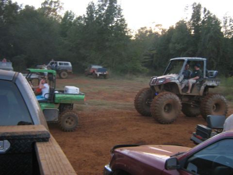 1986 Ford Bronco - Several off-road vehicles are gathered in a muddy field, with trees in the background.