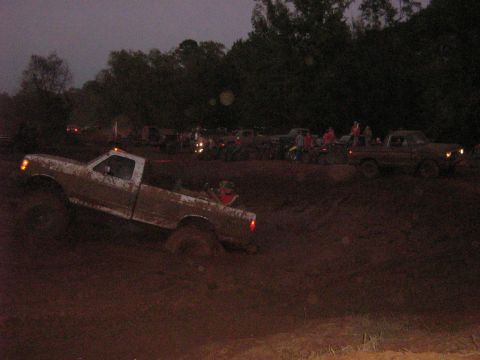 1986 Ford Bronco - A muddy white Ford F-Series truck is stuck in a dirt track with other vehicles in the background.