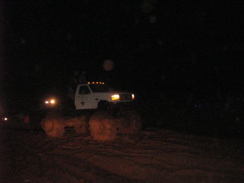 1986 Ford Bronco - A white lifted truck with oversized tires is seen at night, illuminated by its headlights and roof lights.