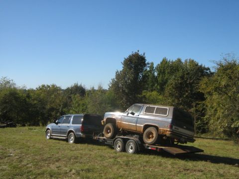 1986 Ford Bronco - A blue SUV tows a trailer carrying a muddy older SUV through a grassy field.