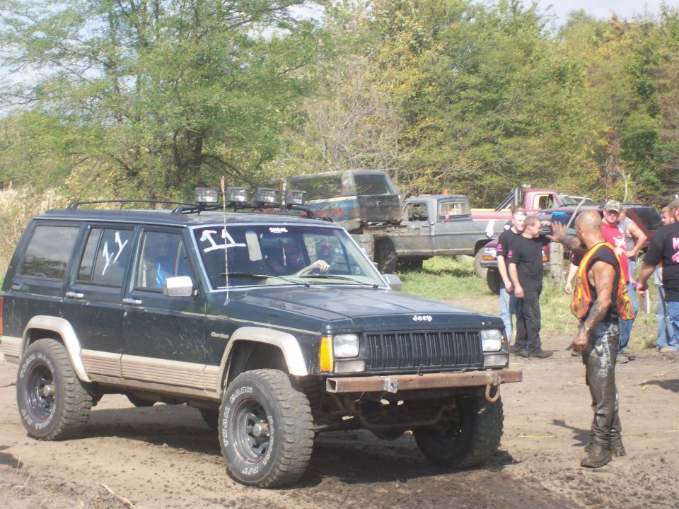 Dark green lifted Jeep Cherokee with roof light bar at a muddy off-road event