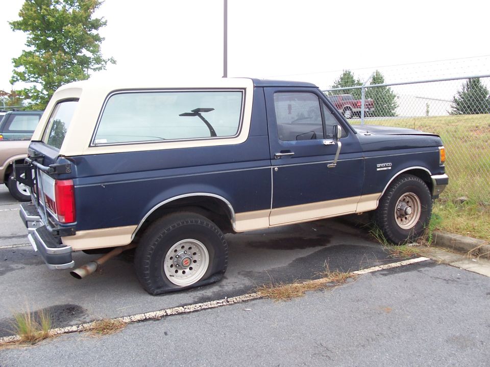 Dark blue and tan two-tone Ford Bronco parked by a chain-link fence with a flat rear tire