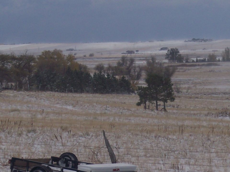trailer in foreground, snow on ground, trees, and distant houses under cloudy sky