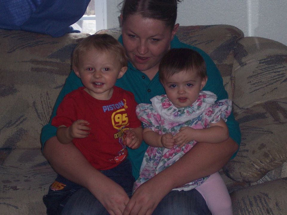 Woman holding a boy in a red shirt with 'PISTON 95' and a baby in a floral dress on a couch