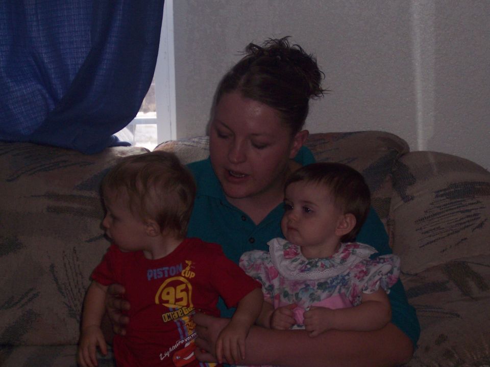 Woman in a teal shirt holds a baby in a floral dress and a toddler in a red shirt with "Piston Cup 95" on it, sitting on a patterned couch