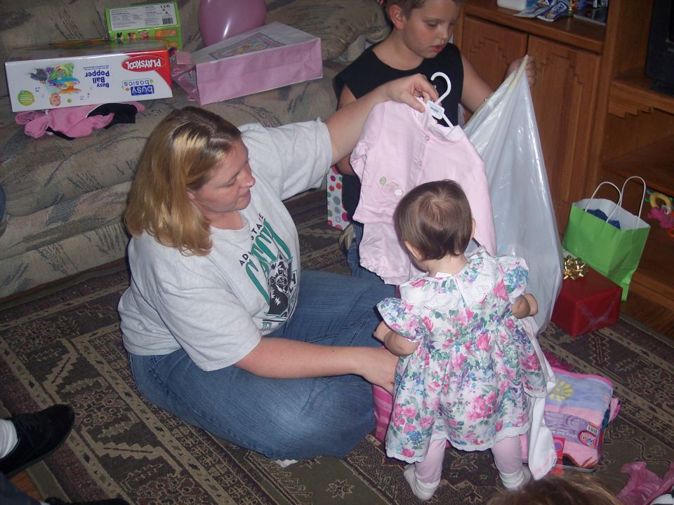 Woman in jeans and t-shirt sits on floor, handing pink shirt to toddler in floral dress. Older child holds shirt on hanger