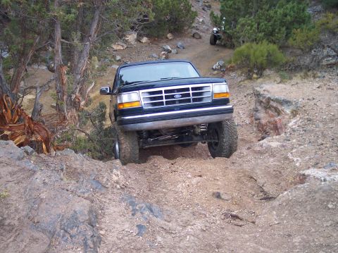 1995 Ford Bronco - A dark blue 1995 Ford Bronco tackles a challenging off-road trail, with another vehicle visible in the background.