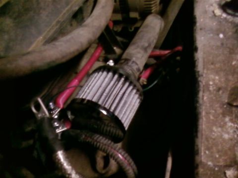 1995 Ford Bronco - Dim engine-bay close-up of a starter motor with its finned solenoid/case, with a heavy red positive battery cable and a green ground wire r…