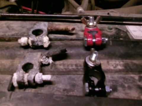 1995 Ford Bronco - Two heavily corroded battery cable terminal clamps, caked in white sulfate buildup, laid on a weathered wooden bench beside their new repla…