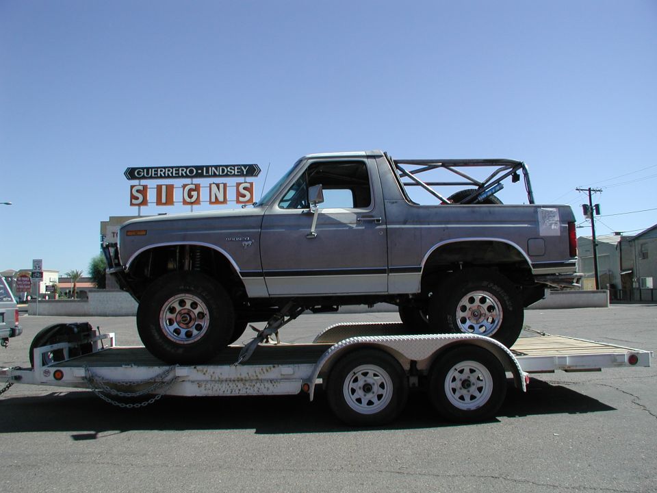 Gray 1980 Ford Bronco on a trailer, lifted with off-road tires, roll cage in the bed