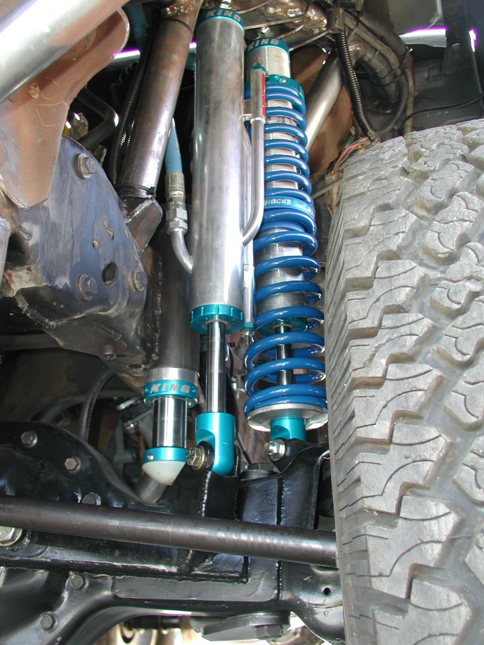 Close-up of blue coilover shocks and silver reservoir shocks on a vehicle's suspension, with a tire visible