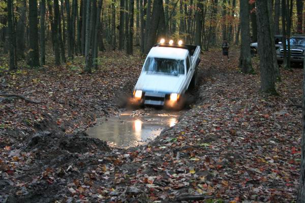 White pickup truck driving through mud, front view. Headlights on, splashing water