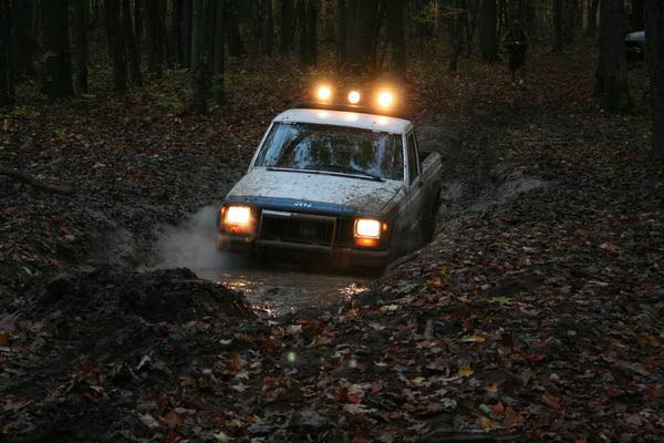 Jeep Comanche driving through a muddy forest path with headlights and roof lights on
