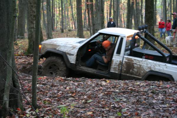White Jeep Comanche pickup truck stuck in mud with driver wearing orange hat. People watch from trees