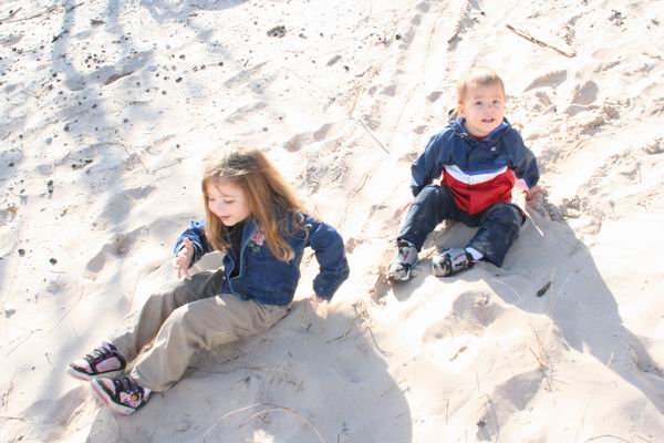 Children sitting on a sand dune, one girl in a denim jacket, one boy in a red, white, and blue jacket. A website URL is visible at the bottom right