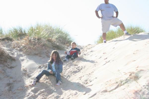Children and adult on sand dunes under a bright sky. A girl sits in the foreground, a boy sits further up the dune, and an adult stands at the top