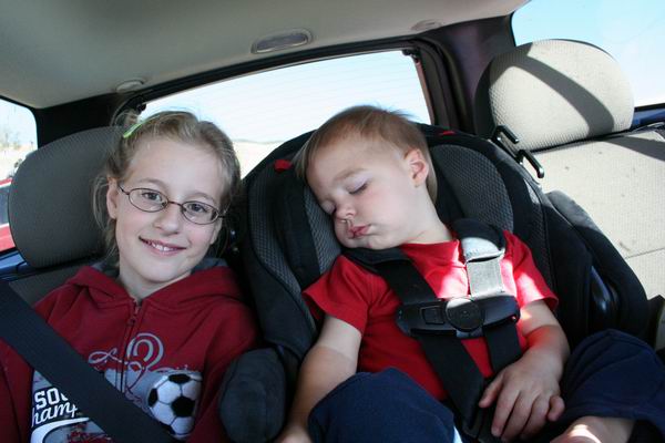 Girl with glasses smiles next to a sleeping toddler in car seats