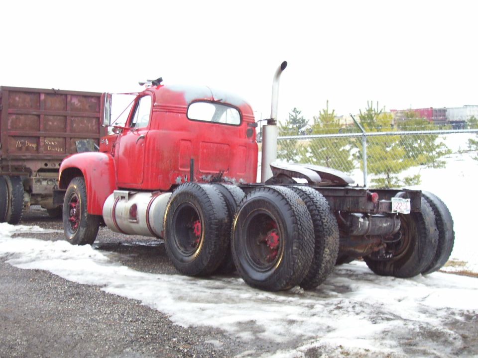 Rear three-quarter view of a red Mack B-series semi tractor with vertical exhaust stack in a snowy lot
