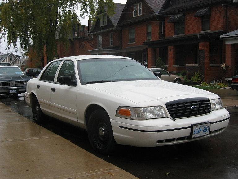 White Crown Victoria sedan with black steel wheels parked on a wet street, front 3/4 view